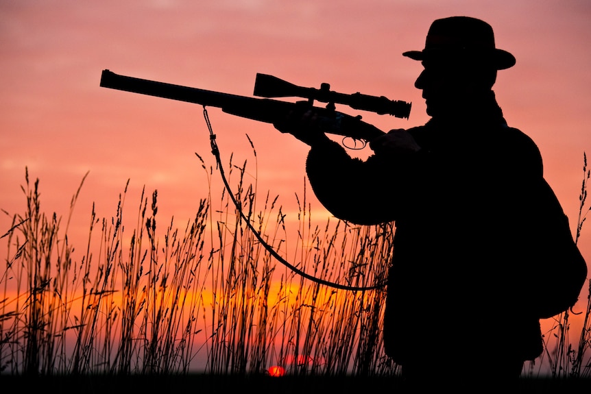 Silhouetts of a hunter aiming his rifle through a telescopic viewfinder as the sun sets behind him.