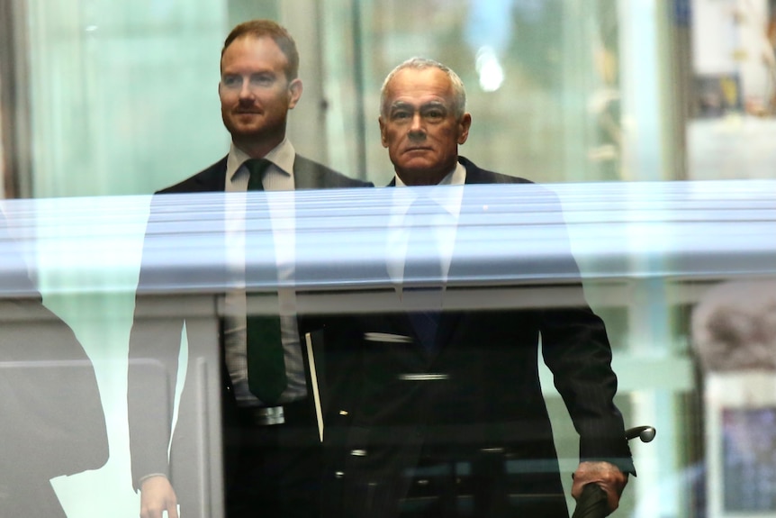 A mid-shot of John Poynton behind glass windows with another man behind to the left.