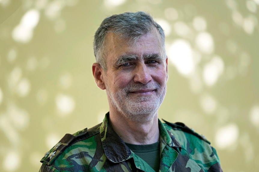 A grey-haired man wearing military fatigues smiles for the camera