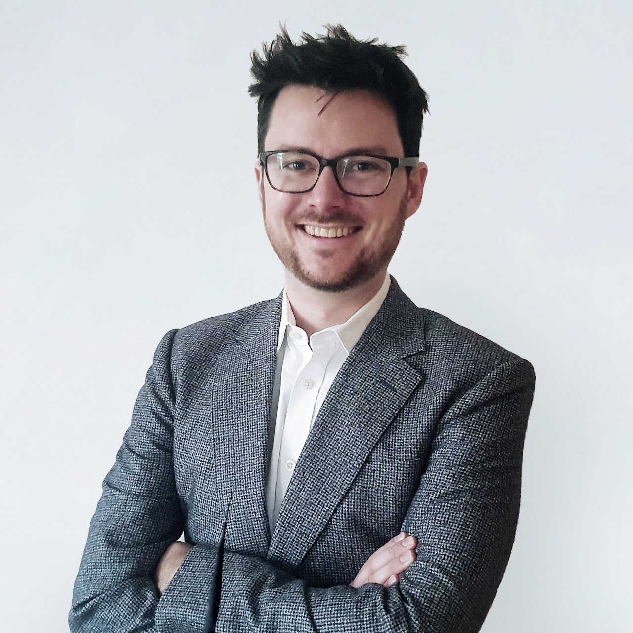 Man with dark hair and glasses smile at camera in front of white background.