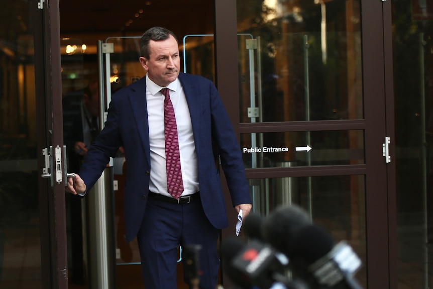 The Premier Mark McGowan wearing a suit with a red tie opens a glass door into a court yard with waiting microphones in the fore