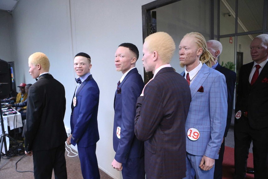 A group of male contestants in suits wait in line in a dimly-lit room during Zimbabwe's 2019 albinism contest.