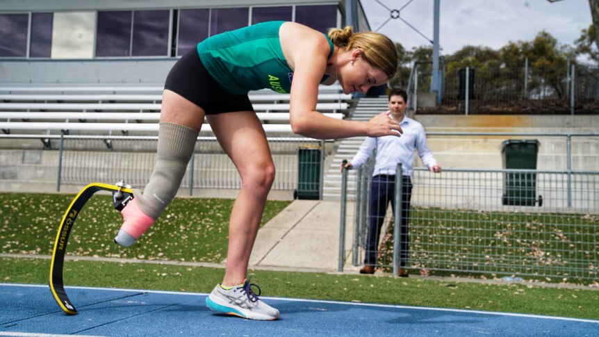 A para-athlete stands in the set position, ready to run with a prosthetic blade on her right leg, with a man watching.