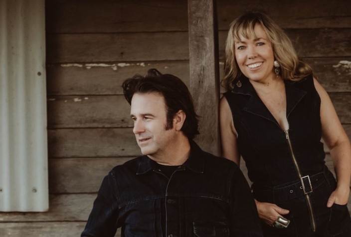 A man and a woman stand in front of a farm house, looking off camera