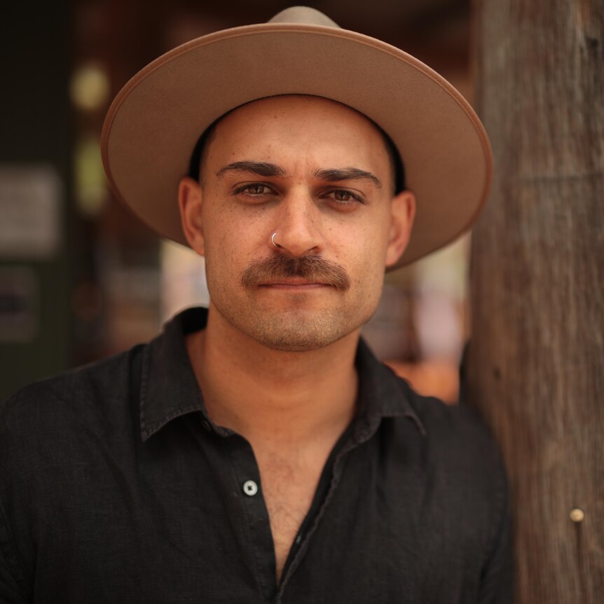 A man with a moustache and nose ring wearing a hat leans against a tree trunk. 