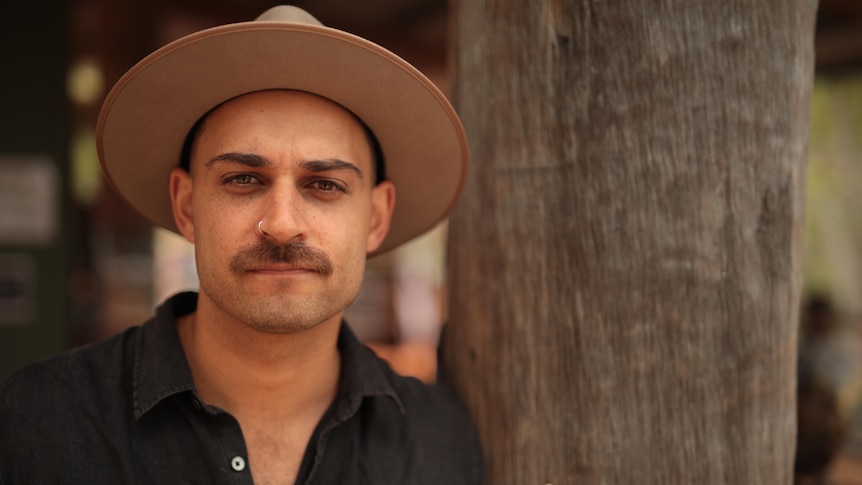 A man with a moustache and nose ring wearing a hat leans against a tree trunk. 