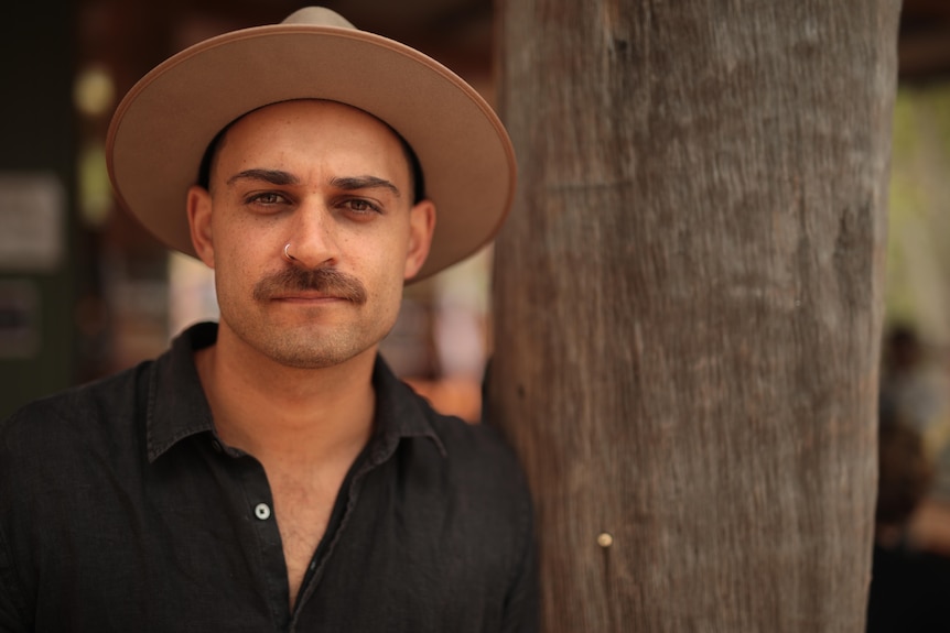 A man with a moustache and nose ring wearing a hat leans against a tree trunk. 