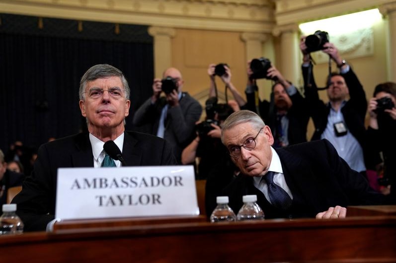 Ambassador Bill Taylor seated in a room, photographers in the background.