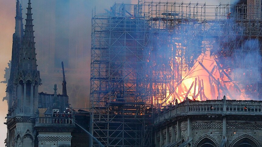 The spire of a gothic cathedral burns along with scaffolding while thick smoke fills the sky