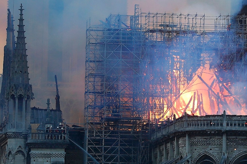 The spire of a gothic cathedral burns along with scaffolding while thick smoke fills the sky