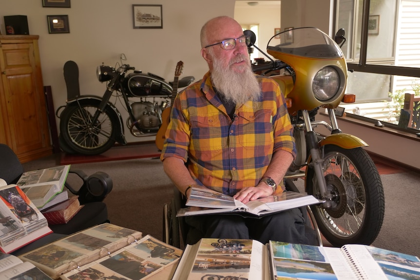 Rob Rees sits inside at a desk looking at photo albums with two motorcycles behind him. 