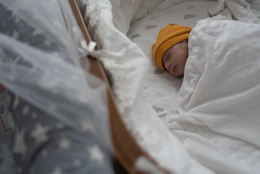 A baby wearing a yellow beanie sleeps in a cot in an orphanage.