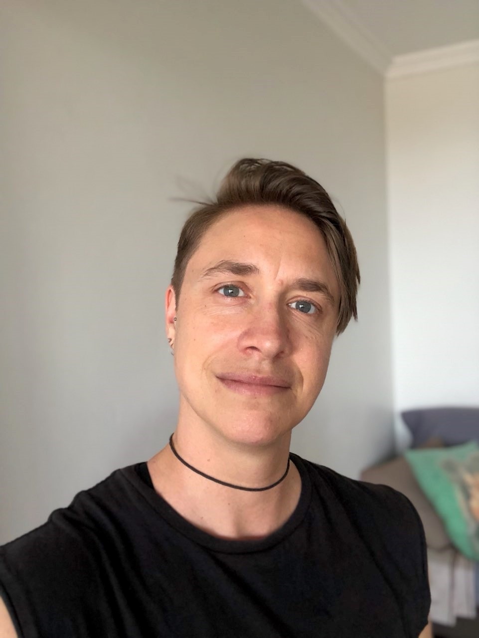 Chris looks to the camera and smiles. They wear a black t-shirt, a black choker necklace and have short hair