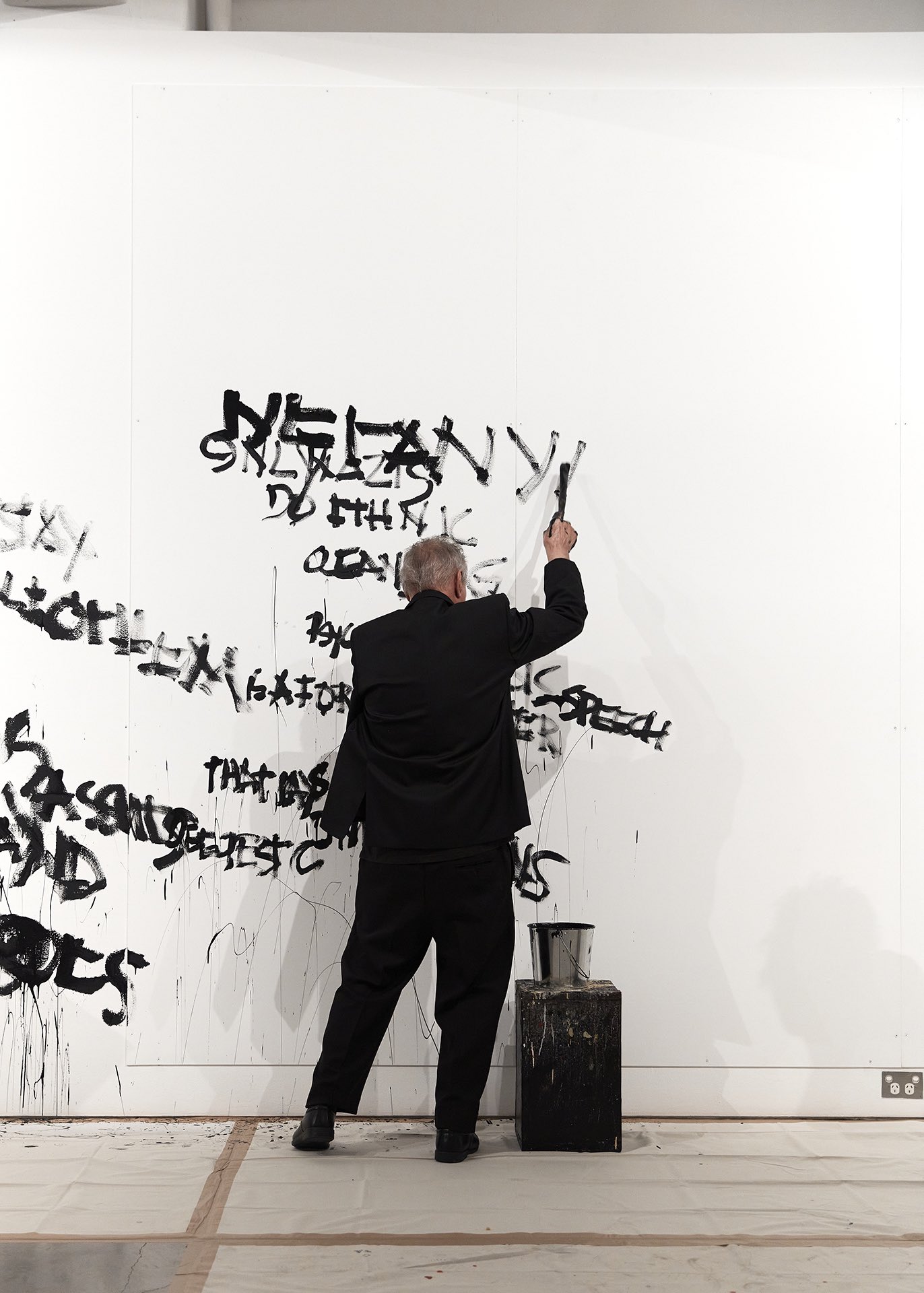 Artist Mike Parr painting text onto a white gallery wall with black paint, while his eyes are closed.