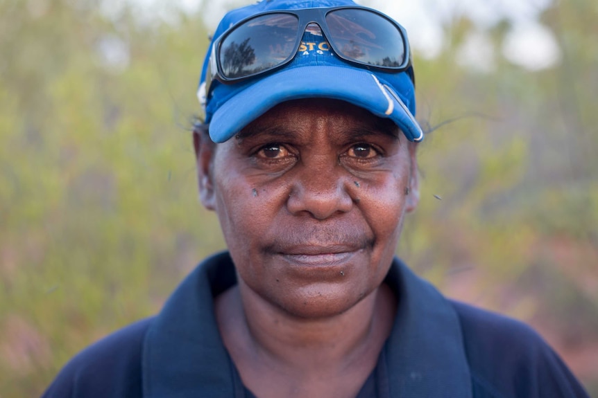 An Indigenous woman wearing a cap with sunglasses perched atop it stares down the barrel of the camera, smiling slightly.
