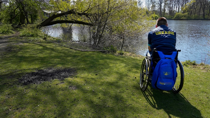 A member of Team Ukraine in a wheechair wearing a team uniform looks out over a lake at the Invictus Games venue in The Hague 