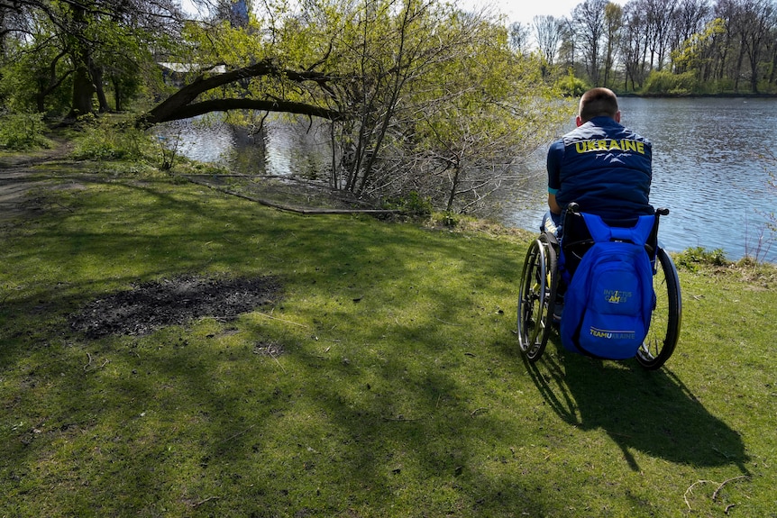 A member of Team Ukraine in a wheechair wearing a team uniform looks out over a lake at the Invictus Games venue in The Hague 
