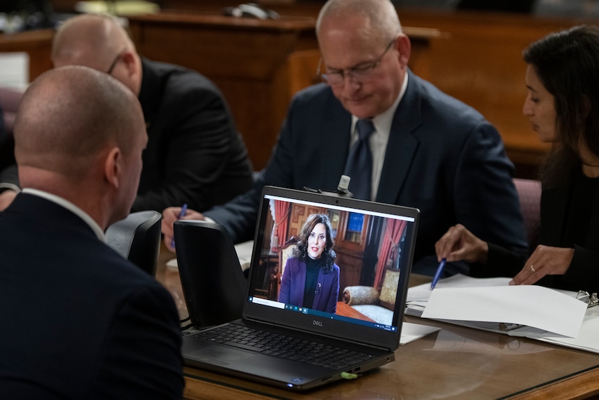 A laptop on a table shows an image of Gretchen Whitmer. Around it are three men and one woman in suits.