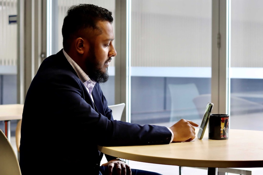 A Bangladeshi man in a suit sits at a table having a video call on his phone.