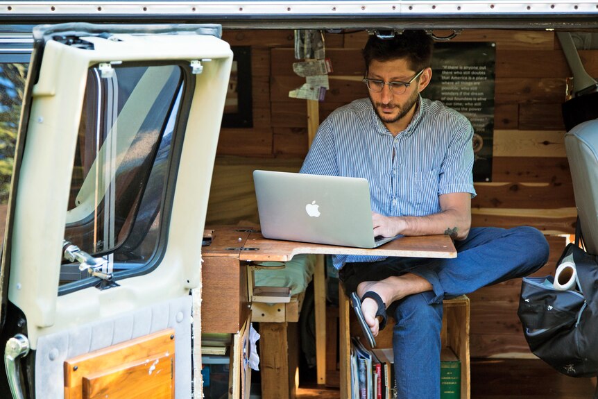 A man wearing glasses works on a laptop out of the back of a van 