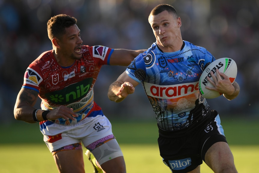 A Cronulla NRL player holds the ball with his left hand as a Newcastle opponent attempts to tackle him.