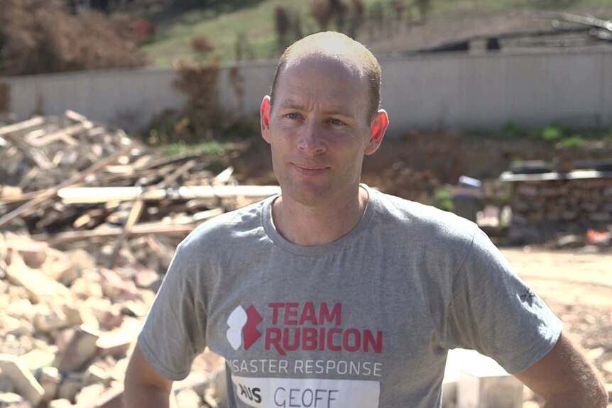 Geoff Evans with grey Team Rubicon t-shirt standing in front of the rubble of a house destroyed in a bushfire