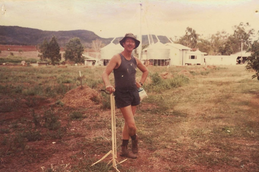 An older image of a blue singlet-wearing pig farmer standing in a paddock with sheds and silos in the background