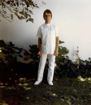 A young man, smiling, stands by a white wall in a garden, wearing white nurse's scrubs.