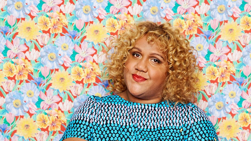 Elaine Crombie with curly blonde hair, gently smiles while posing in front of a floral wall.