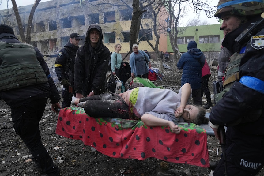 A group of men use a stretcher to carry an injured pregnant woman through rubble outside of a damaged hospital building.