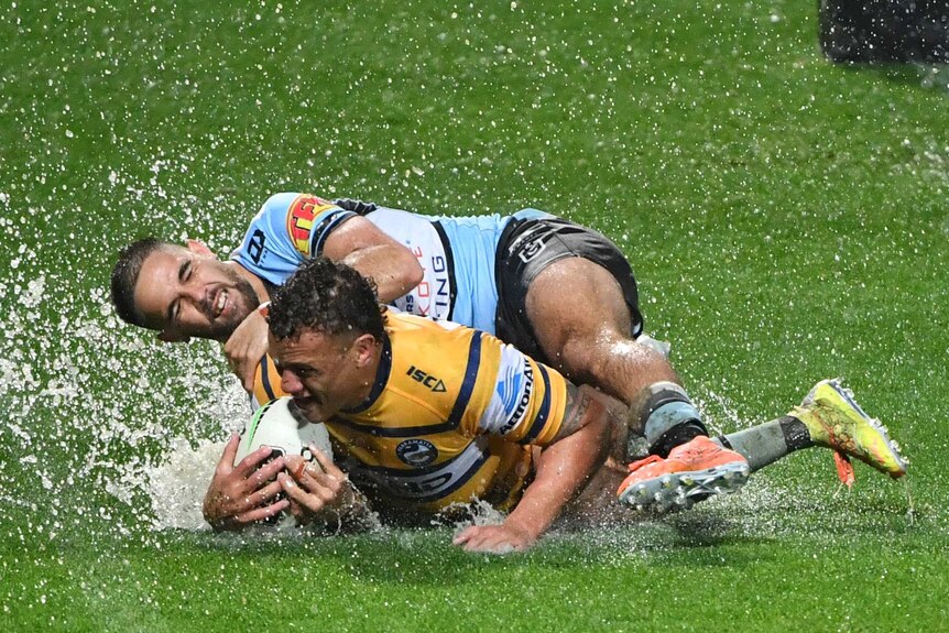 A Parramatta Eels NRL player scores a try with a Cronulla opponent tackling him as water spurts up from the grass.