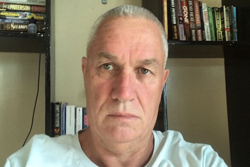 Graeme Cahill looks to the camera with a neutral expression on his face. He sits in front of a black bookshelf.