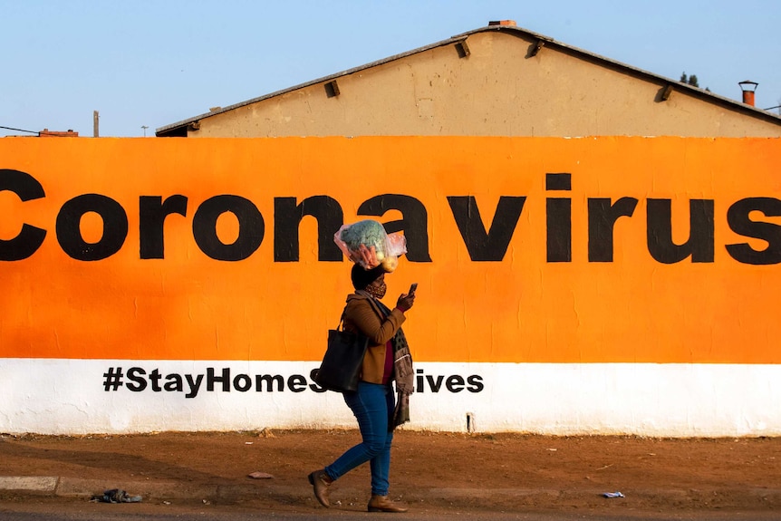 A woman carrying groceries on her head waring a face mask walks past a billboard with a coronavirus health warning.