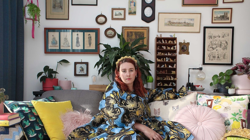 Emma Batchelor wears a floral blue dress and is sitting on a couch in her living room. She is looking directly at the camera.