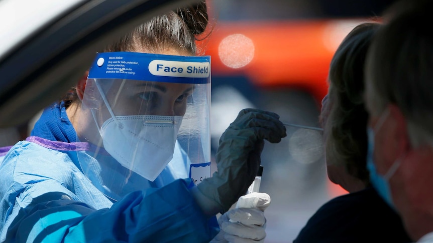 Woman in protective equipment and face shield uses a swab on a patient in a car.