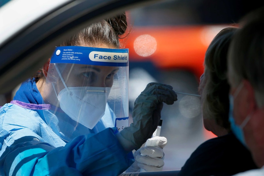 Woman in protective equipment and face shield uses a swab on a patient in a car.
