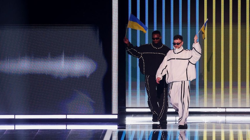 Two people one a black tracksuit, one in a white tracksuit walk out holding Ukrainian flags