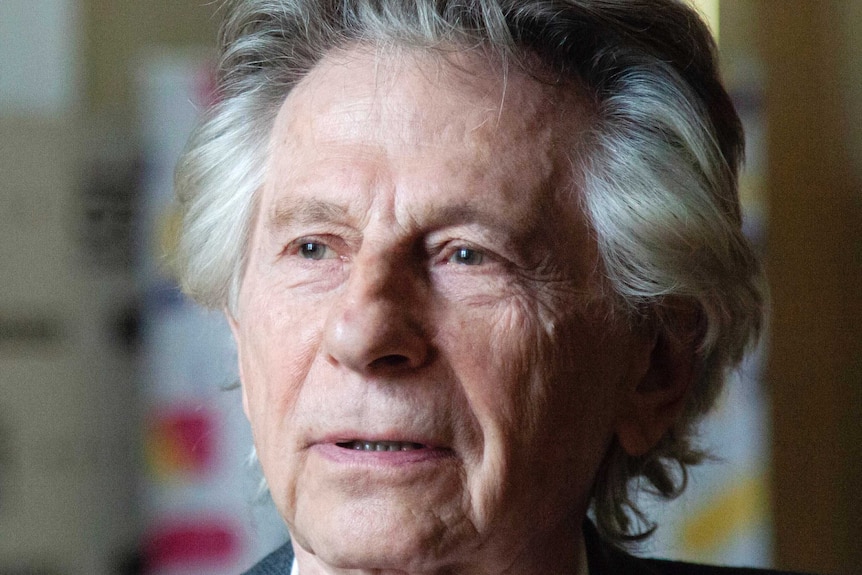 A close up shot of a grey-haired elderly man, wearing a suit, in a naturally lit room.