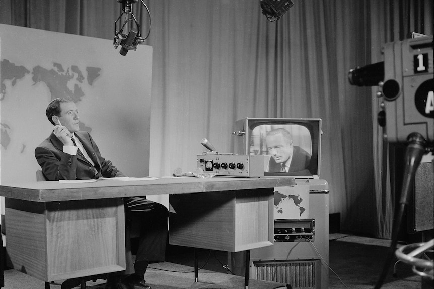 Black and white photo of old TV studio with man sitting at a desk smoking.