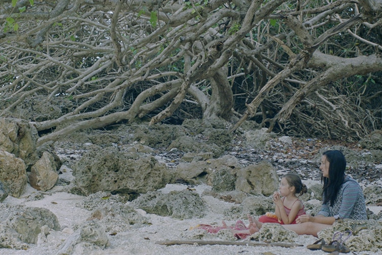 Colour still of Poh Lin Lee sitting with daughter on beach in 2018 film Island of the Hungry Ghosts.