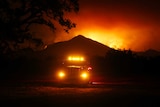A truck with glowing orange headlights sits in front of a burning hill.