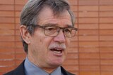 A head and shoulders shot of Mike Nahan speaking to reporters in front of a brick wall.