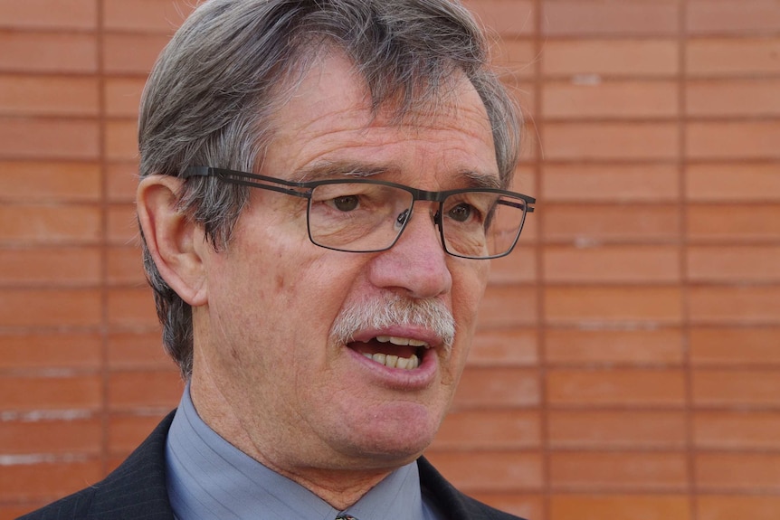 A head and shoulders shot of Mike Nahan speaking to reporters in front of a brick wall.