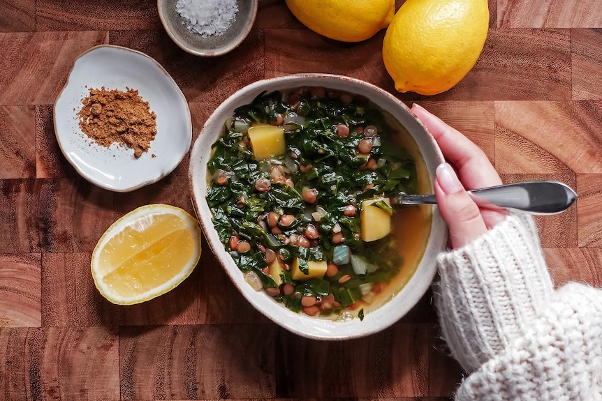 A hand hugging a bowl of lentil soup with potato and spinach. On the table are lemons, cumin and sea salt.