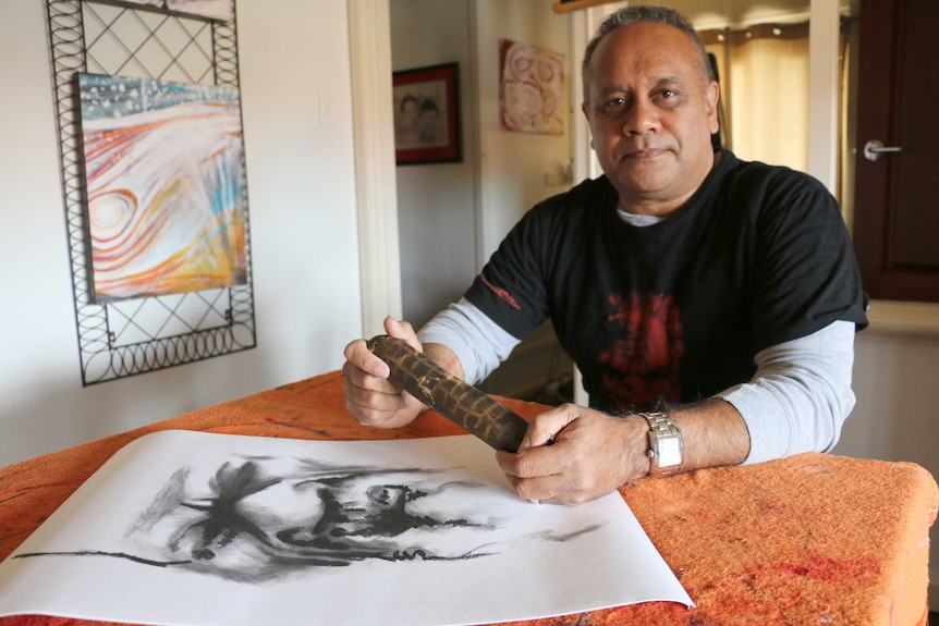 Barry McGuire sits at a table indoors holding a wooden item with a black and white painting on the table.