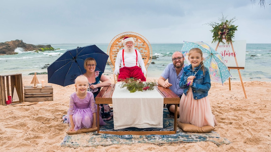 A mother and father sit with their daughters next to Santa on a beach, holding umbrellas.