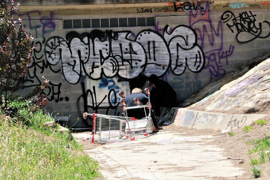 Two teenagers sit next to a graffiti wall, with one bent over as if smoking something.