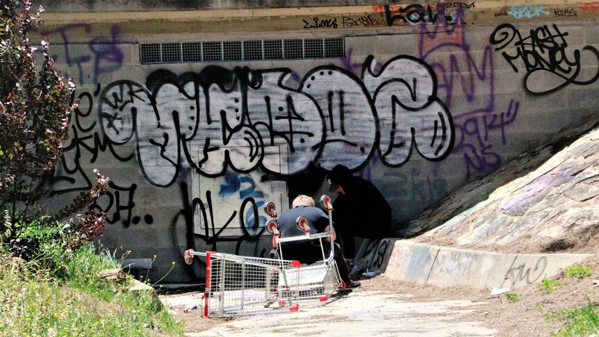Two teenagers sit next to a graffiti wall, with one bent over as if smoking something.