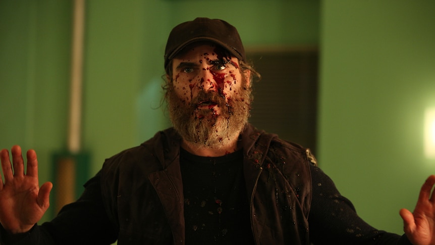Joaquin Phoenix dressed in black with blood splatter on his face and both hands raised in film You Were Never Really Here.