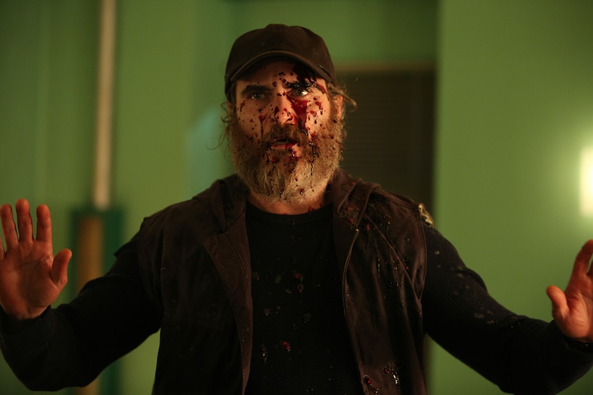 Joaquin Phoenix dressed in black with blood splatter on his face and both hands raised in film You Were Never Really Here.